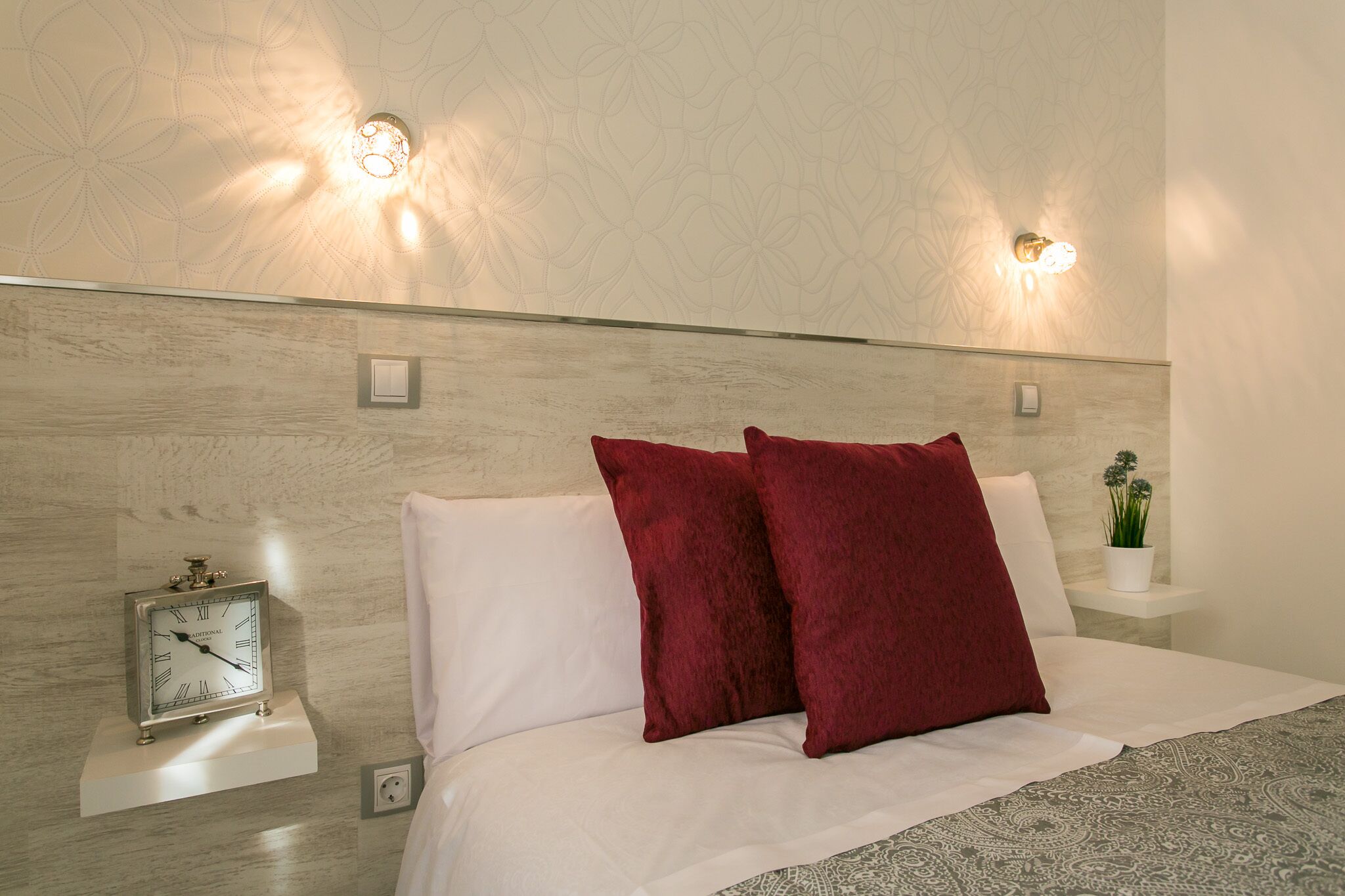 A modern bedroom with a bed, two red pillows, a wall clock, and two wall-mounted lights.
