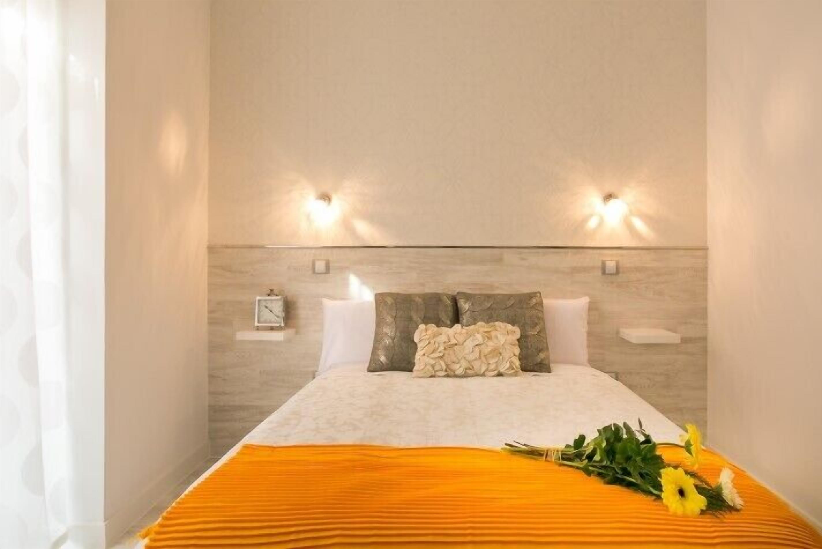Una cama bien hecha con edredón blanco, dos almohadas y una almohada decorativa. Hay dos lámparas de pared a cada lado de la cama.