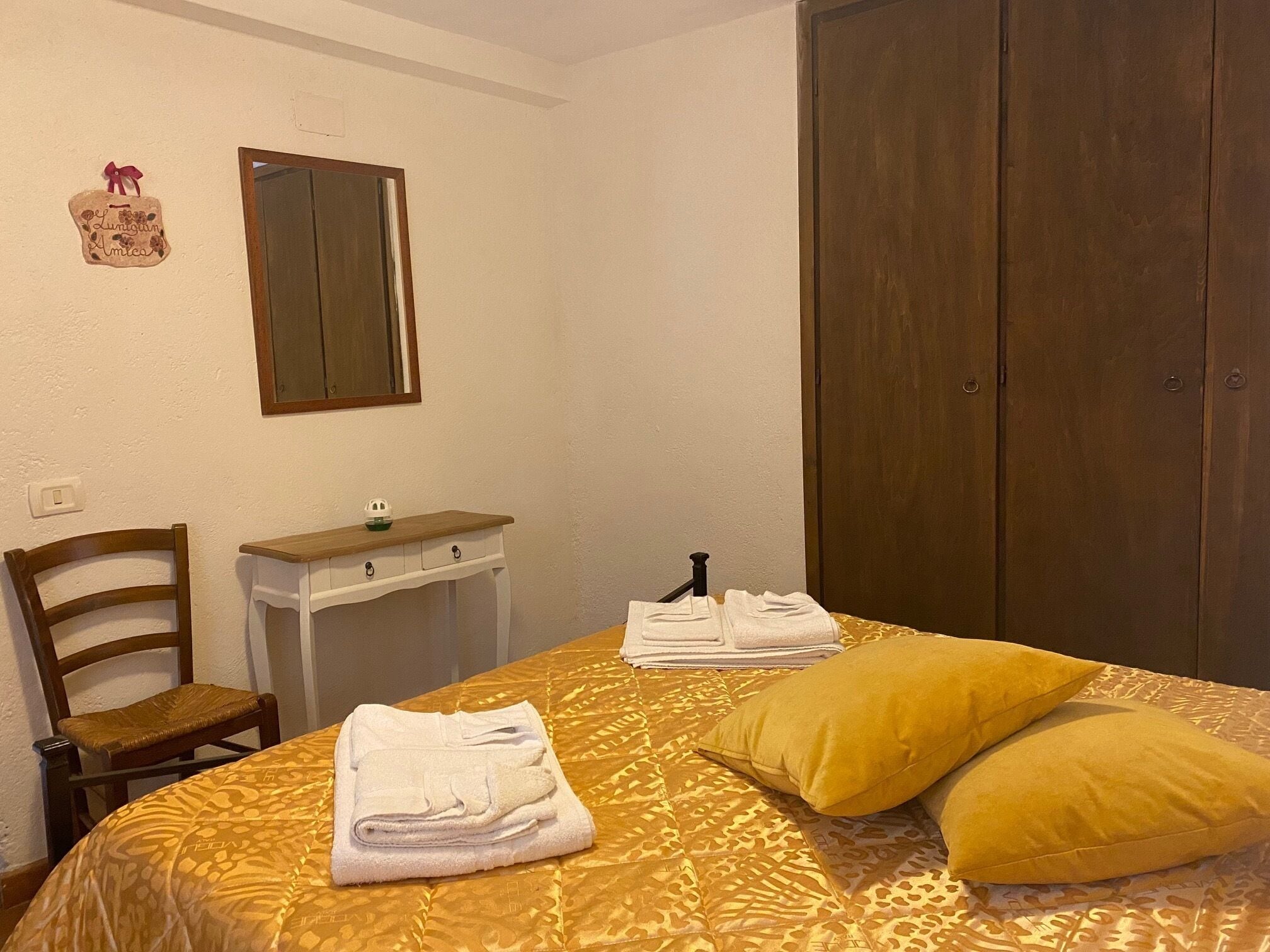 Quarto de hotel com cama, uma cadeira de madeira, uma mesinha com espelho e um guarda-roupa.