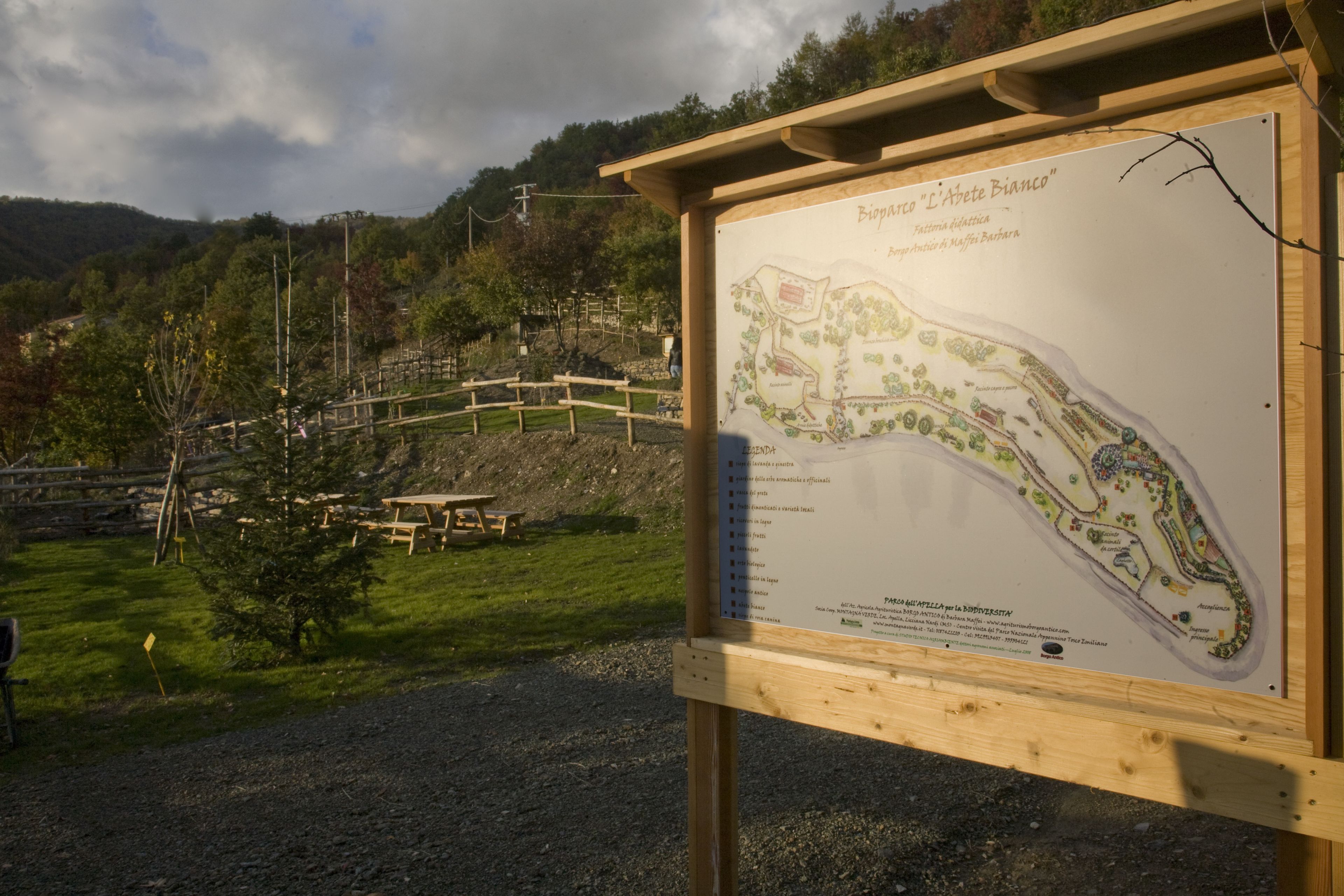 A wooden signboard with a map and text, set against a backdrop of a park with a wooden fence, picnic tables, and trees.