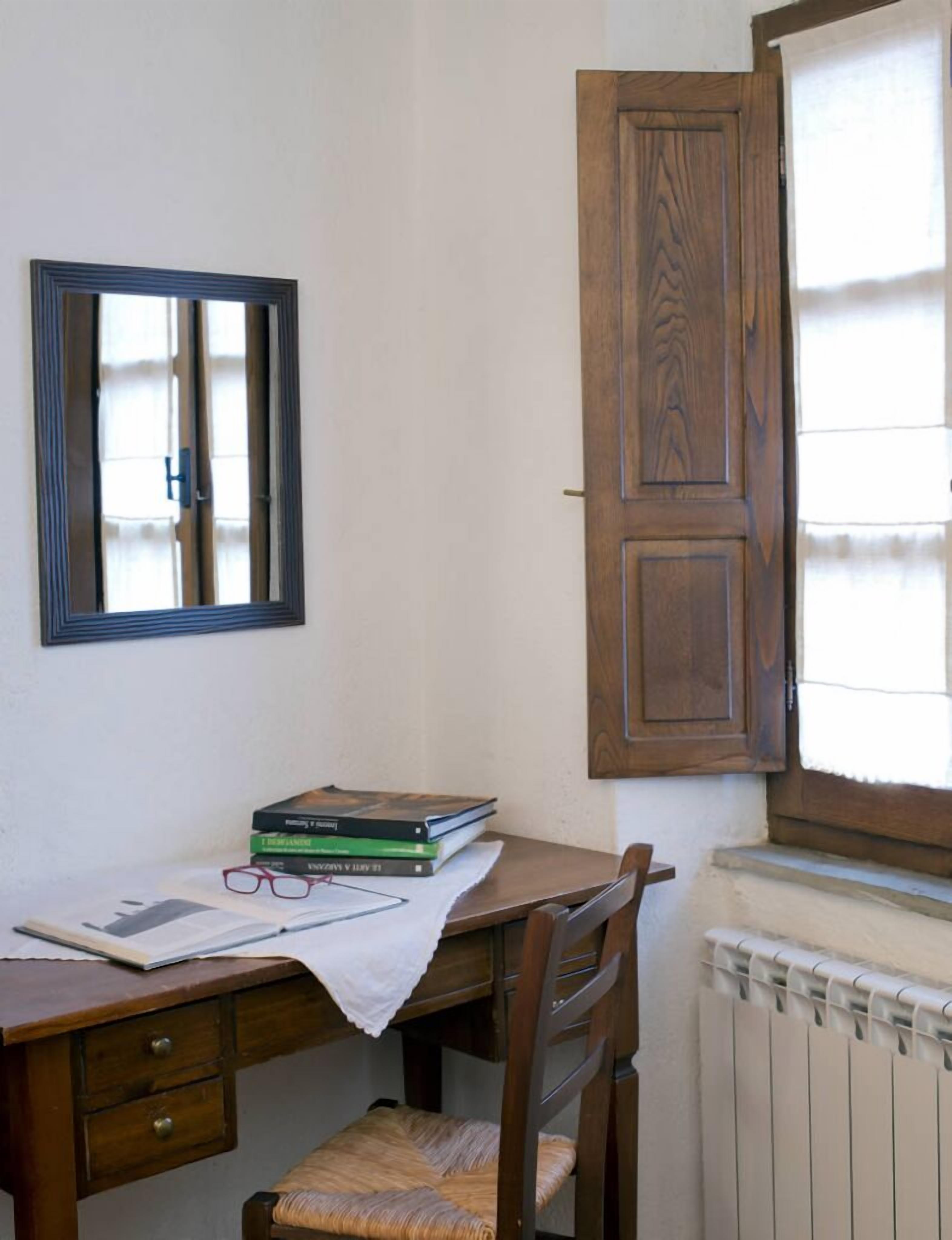 Une salle d’étude avec un bureau en bois, des livres, un miroir et une fenêtre avec des volets en bois.