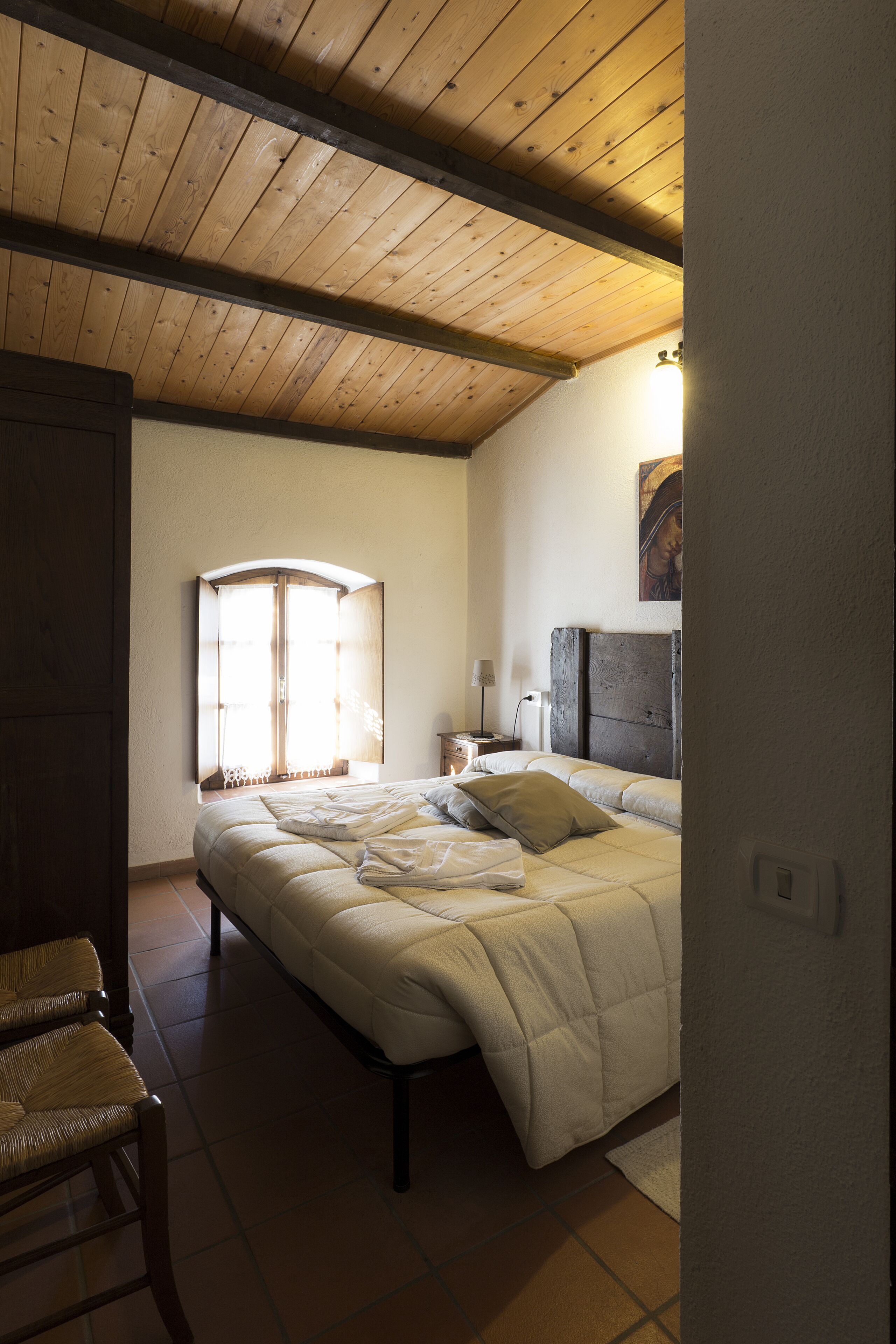 Una camera da letto con soffitto in legno, un letto con testiera, una finestra con tende e un comodino con una lampada.