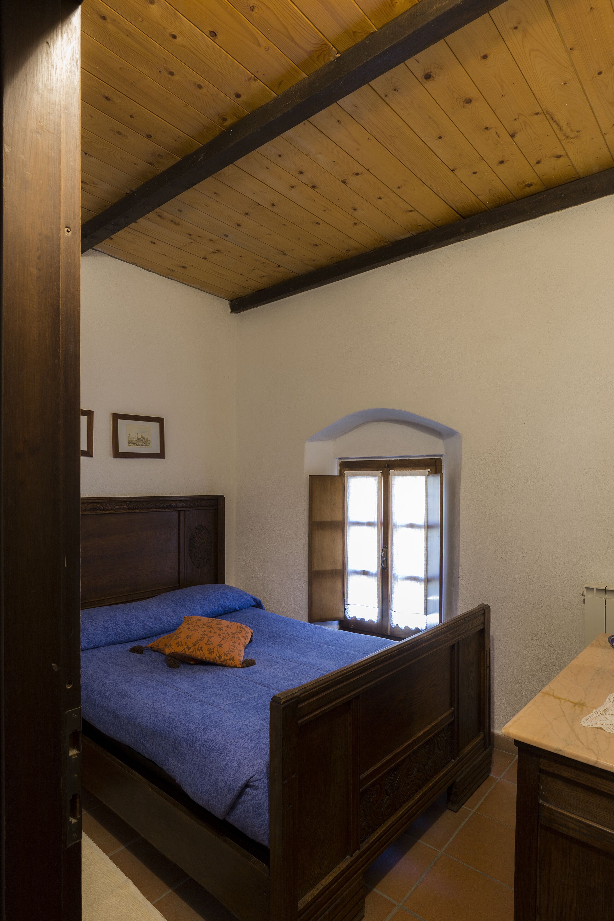 Camera da letto con soffitto in legno, un letto con copriletto blu, una testiera in legno e una finestra con cornice bianca.