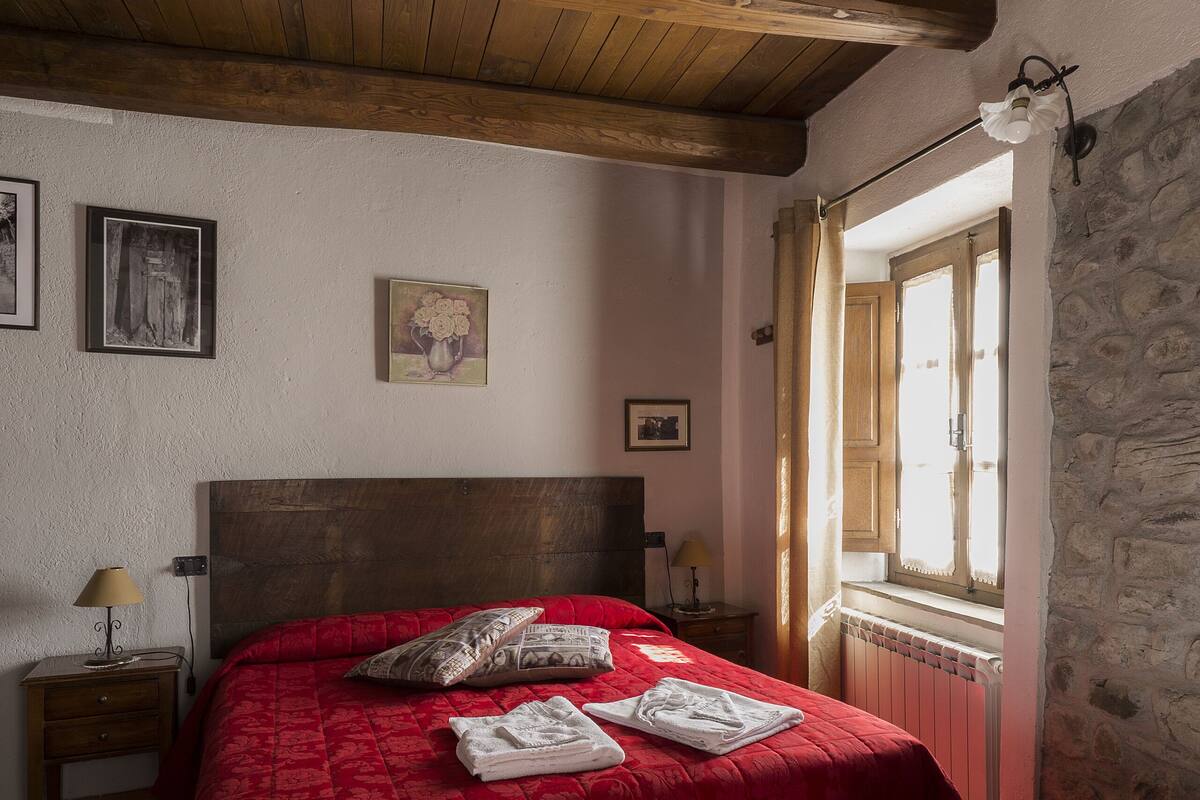 Ein Schlafzimmer mit Holzbalkendecke, einem großen Bett mit roter Bettwäsche, einem Nachttisch mit Lampe und einer Steinwand.