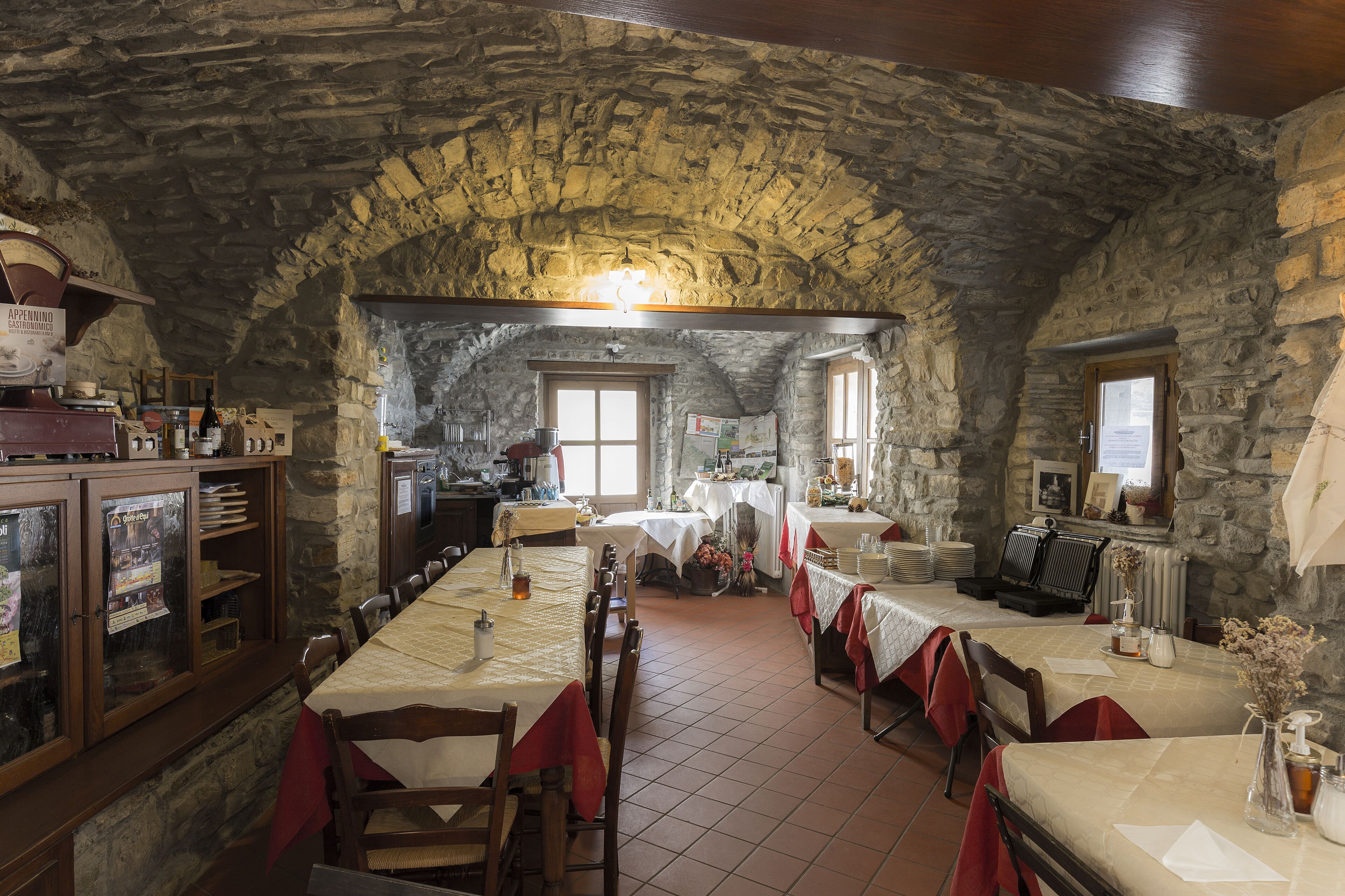 A rustic stone-walled dining area with wooden tables and chairs, a kitchen in the background, and a stone ceiling.
