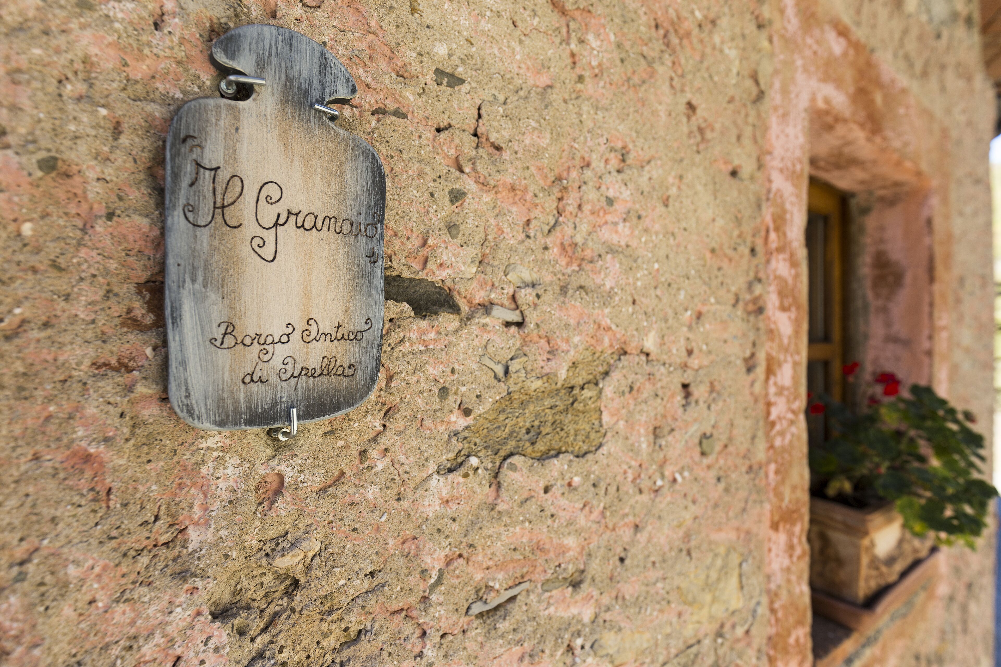 Une enseigne en bois portant l’inscription « Il Granoio Borgo Cinese di Spelza » est fixée à un mur de pierre.