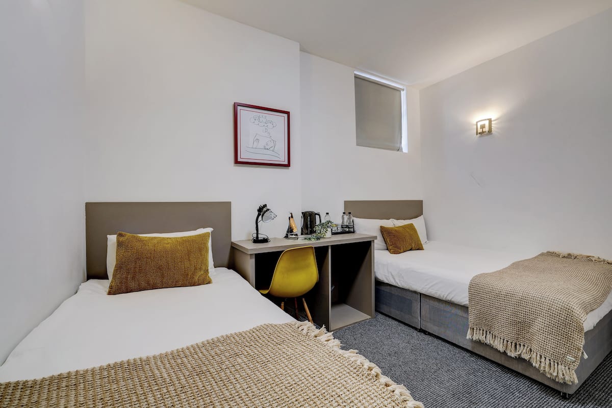 Une chambre d’hôtel avec deux lits, un bureau, une chaise, une petite table, une lampe fixée au mur et une fenêtre.