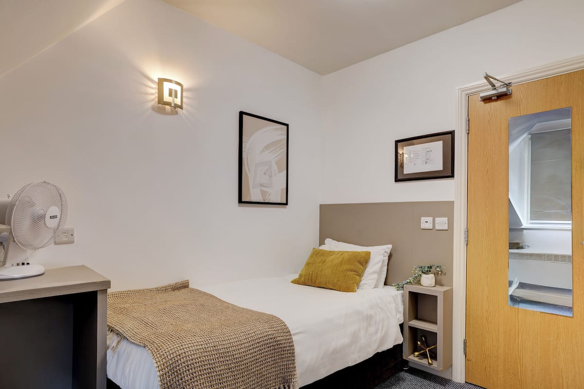 Une chambre d’hôtel bien rangée, avec un lit, une table de chevet, un ventilateur et une lampe fixée au mur.
