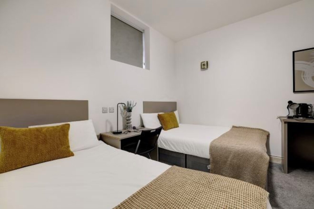 Une chambre d’hôtel avec deux lits, un bureau, une chaise, une petite table avec une lampe et une fenêtre.