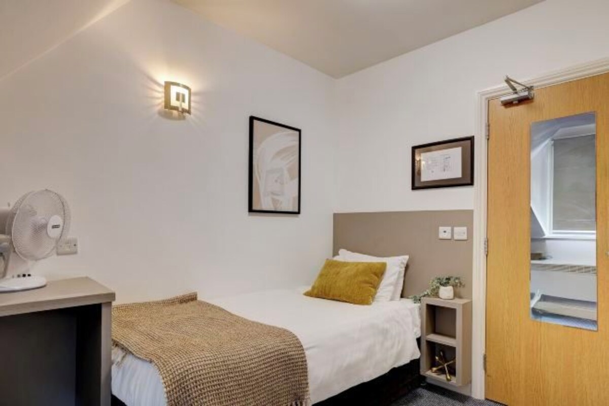 Une chambre à coucher comprenant un lit, une table de chevet, un ventilateur fixé au mur et une porte avec une fenêtre.