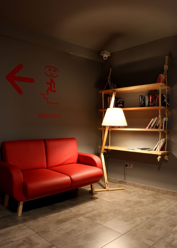 A red sofa, a wooden bookshelf, and a wall-mounted lamp in a room with tiled flooring.