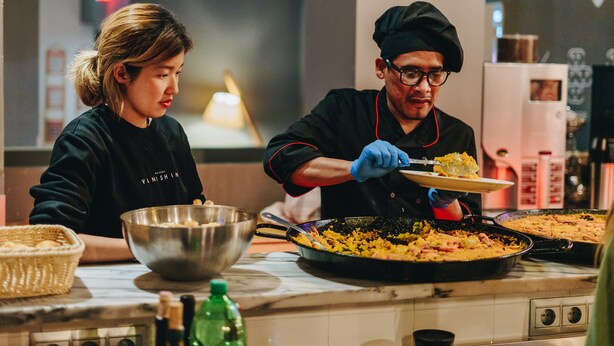 A kitchen with two people cooking, a large pan on the stove, and a bowl on the counter. - Ok Hostel Madrid (Madrid)