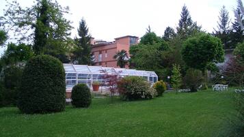 Ein Gewächshaus mit Glasdach, umgeben von gepflegten Rasenflächen und verschiedenen Bäumen.