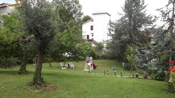 Uma área ao ar livre com gramado, um parquinho infantil com escorregador e balanços, cercada por árvores e um prédio branco ao fundo.