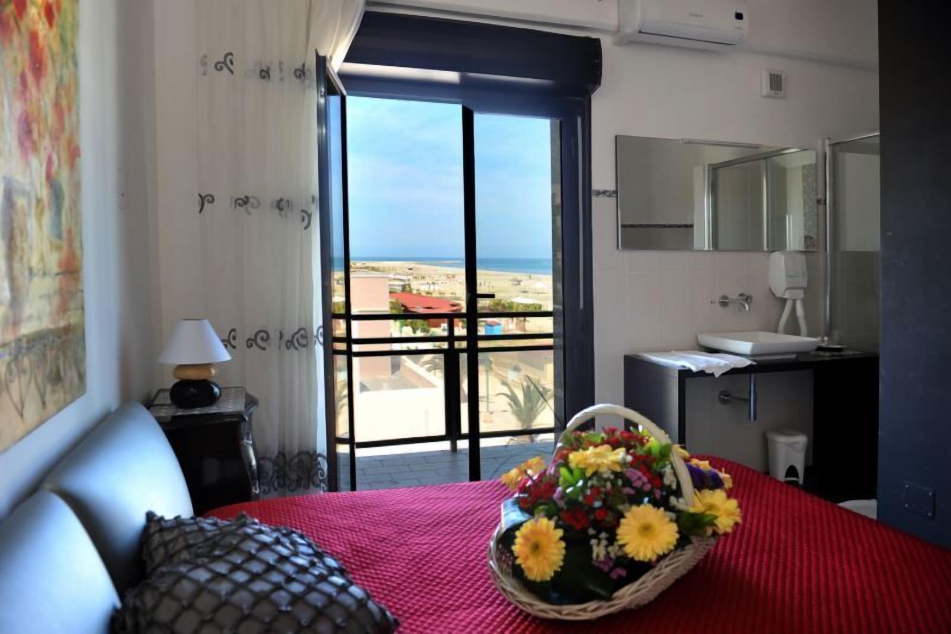 Una habitación de hotel con una cama, un ventanal con vista a la playa, un baño y una mesita de noche con una lámpara y un cesto de flores.