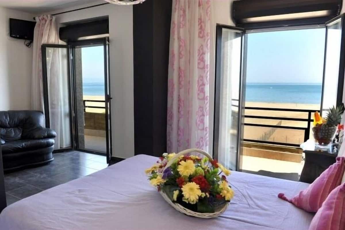 Ein Hotelzimmer mit Bett, Sessel, Fenster mit Blick auf den Strand und einer Blumenkorb.