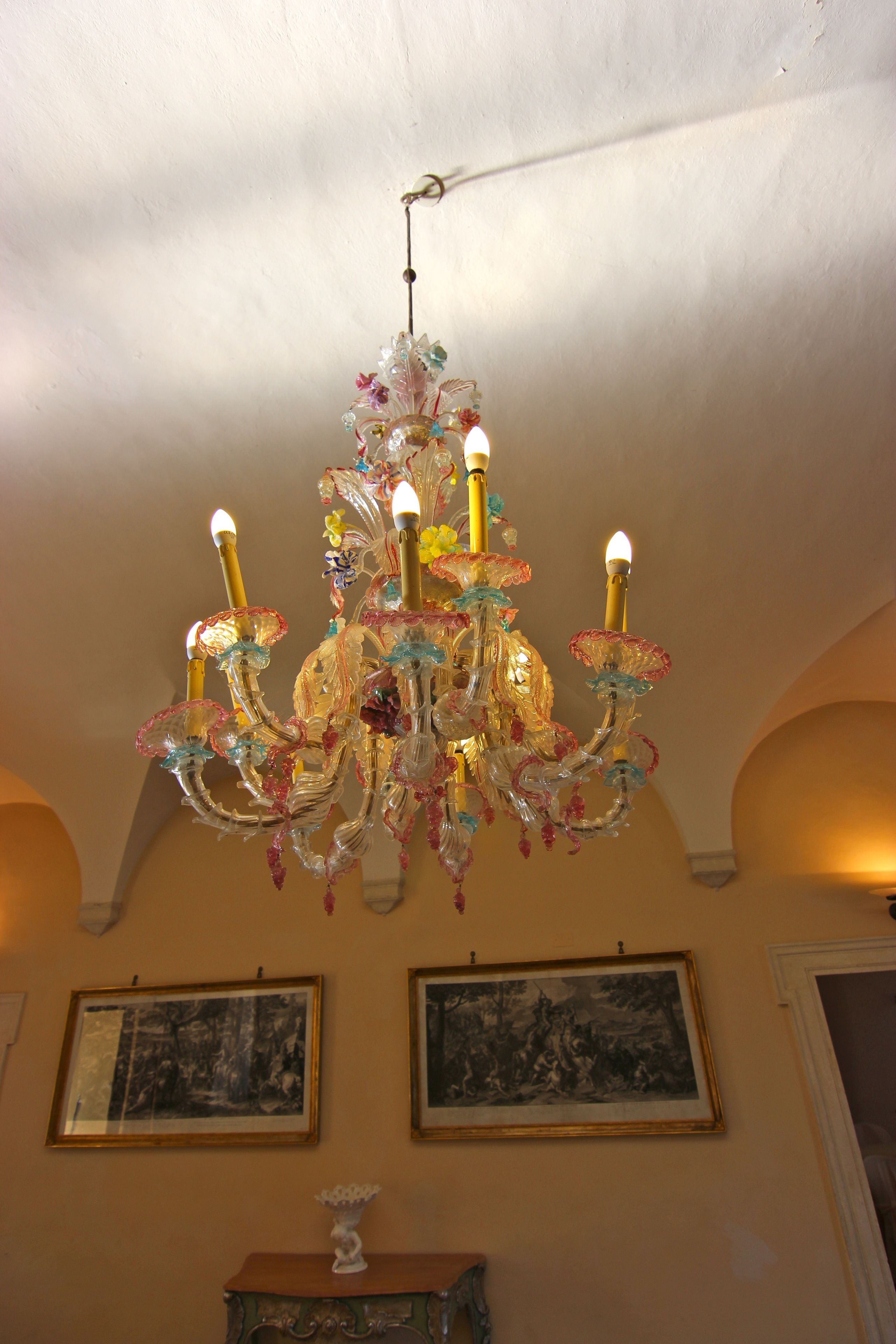 Ein Kronleuchter mit mehreren Armen und Lichtern, der an der Decke in einem Raum hängt, der zwei gerahmte Bilder zeigt.