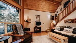 A cozy living room with a fireplace, a sofa with a 'RELAX' pillow, a wooden staircase, and a large window overlooking trees. - 04sl- Hot Tub - Bbq - Pets Ok - Sleeps 5 1 Bedroom Home by RedAwning (Deming)