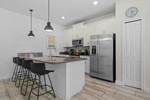 A modern kitchen with white cabinets, a stainless steel refrigerator, a microwave, and a coffee maker. There are three black bar stools and pendant lights above the island. - Champions Gate Resort (Davenport)