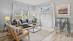 Un salon moderne avec un canapé gris, un fauteuil en bois et une table basse. Il y a deux fenêtres avec des rideaux blancs, une lampe et un tableau encadré au mur.