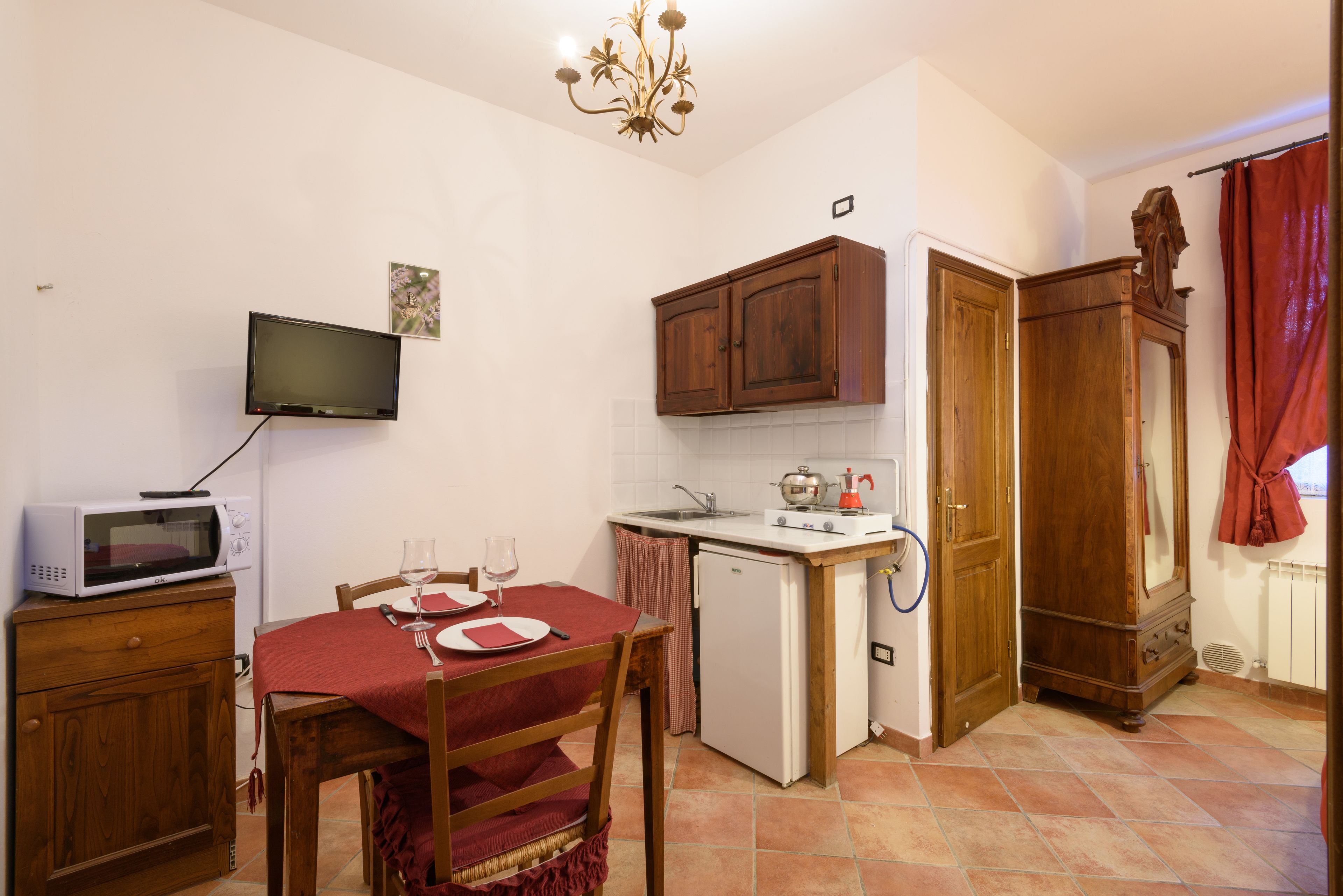 A compact kitchen with a microwave, a small refrigerator, wooden cabinets, a dining table with chairs, and a television mounted on the wall.