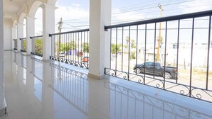 Un pasillo con columnas blancas y suelo reflectante, que conduce a un balcón con baranda metálica y vistas a una calle con coches estacionados.