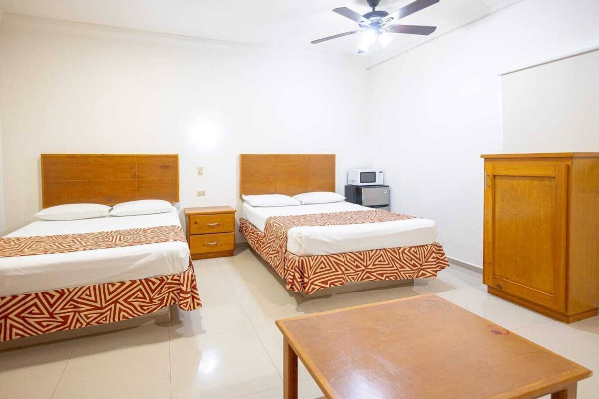 Habitación de hotel con dos camas, cabecero de madera, mesita de noche pequeña, armario y ventilador de techo.