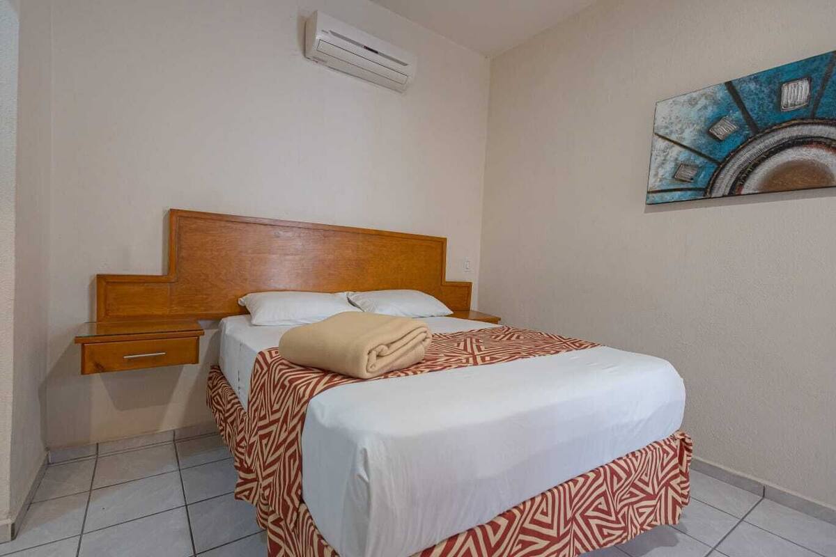 Habitación de hotel con una cama de cabecera de madera, una mesita de noche, una lámpara de noche y un cuadro en la pared.