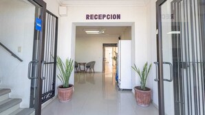 Una zona de recepción con un cartel sobre la puerta, dos plantas en macetas y un área de estar al fondo.