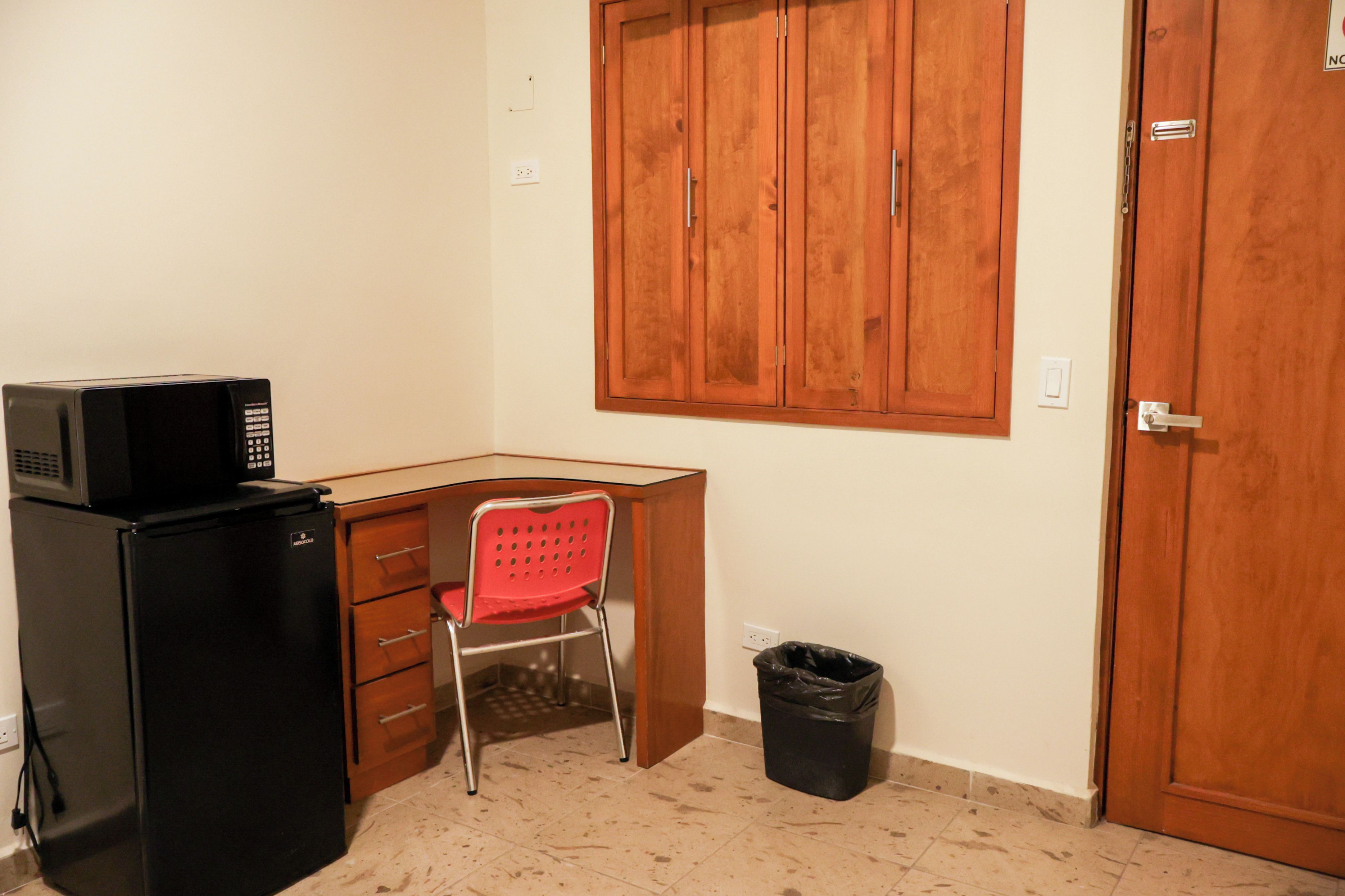 Una habitaci ón pequeña con un escritorio de madera, una silla roja, un cesto de basura negro y una puerta de madera cerrada.