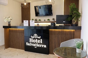Un mostrador de recepción de hotel con un televisor de pantalla plana montado encima, una planta en maceta a la derecha y un letrero que dice 'Hotel Salvatierra'.
