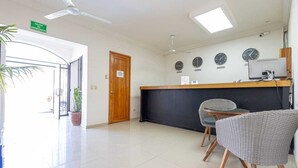 Un área de recepción con mostrador, dos sillas de mimbre, un ventilador de techo y un reloj en la pared.