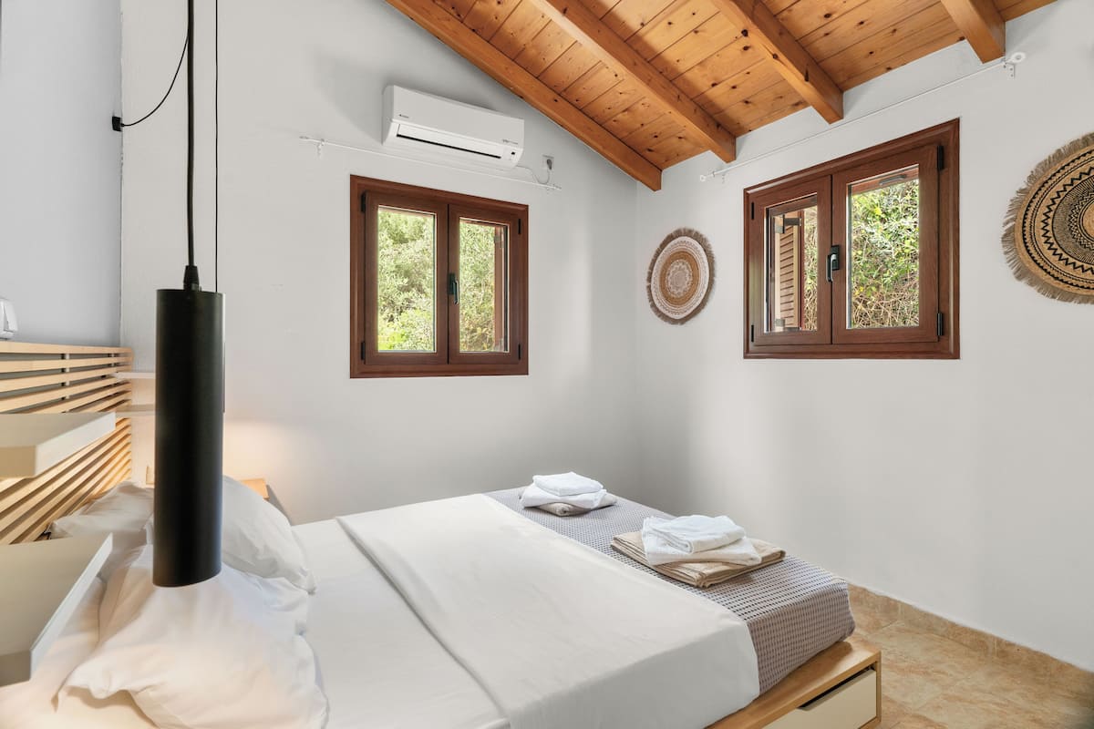 Ein Schlafzimmer mit einem Bett, einem Nachttisch, einer Hängelampe, zwei Fenstern und einer Wand-Klimaanlage.