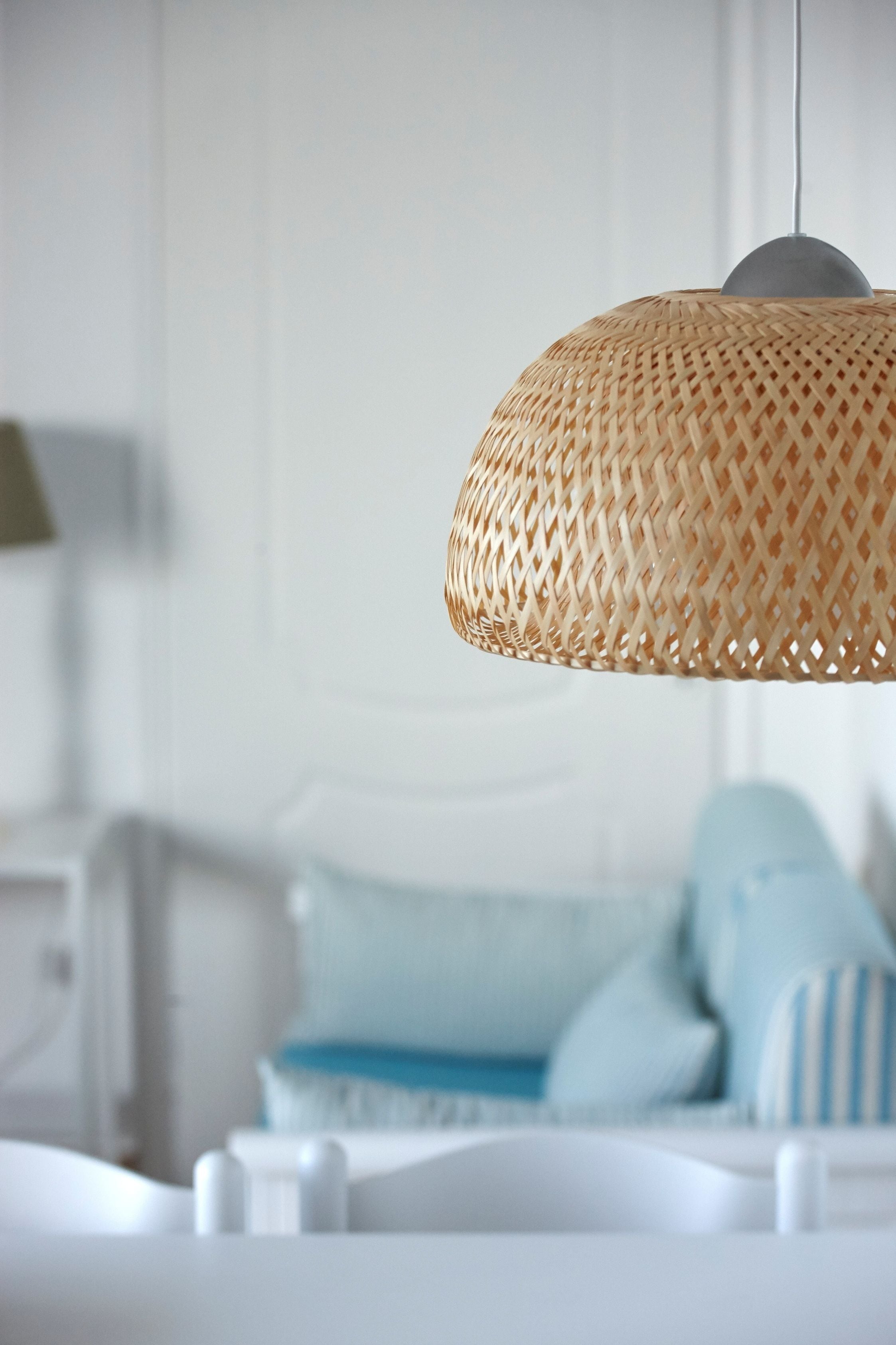 Un luminaire suspendu en osier dans une pièce où se trouve un lit blanc et un coussin à rayures bleues et blanches.
