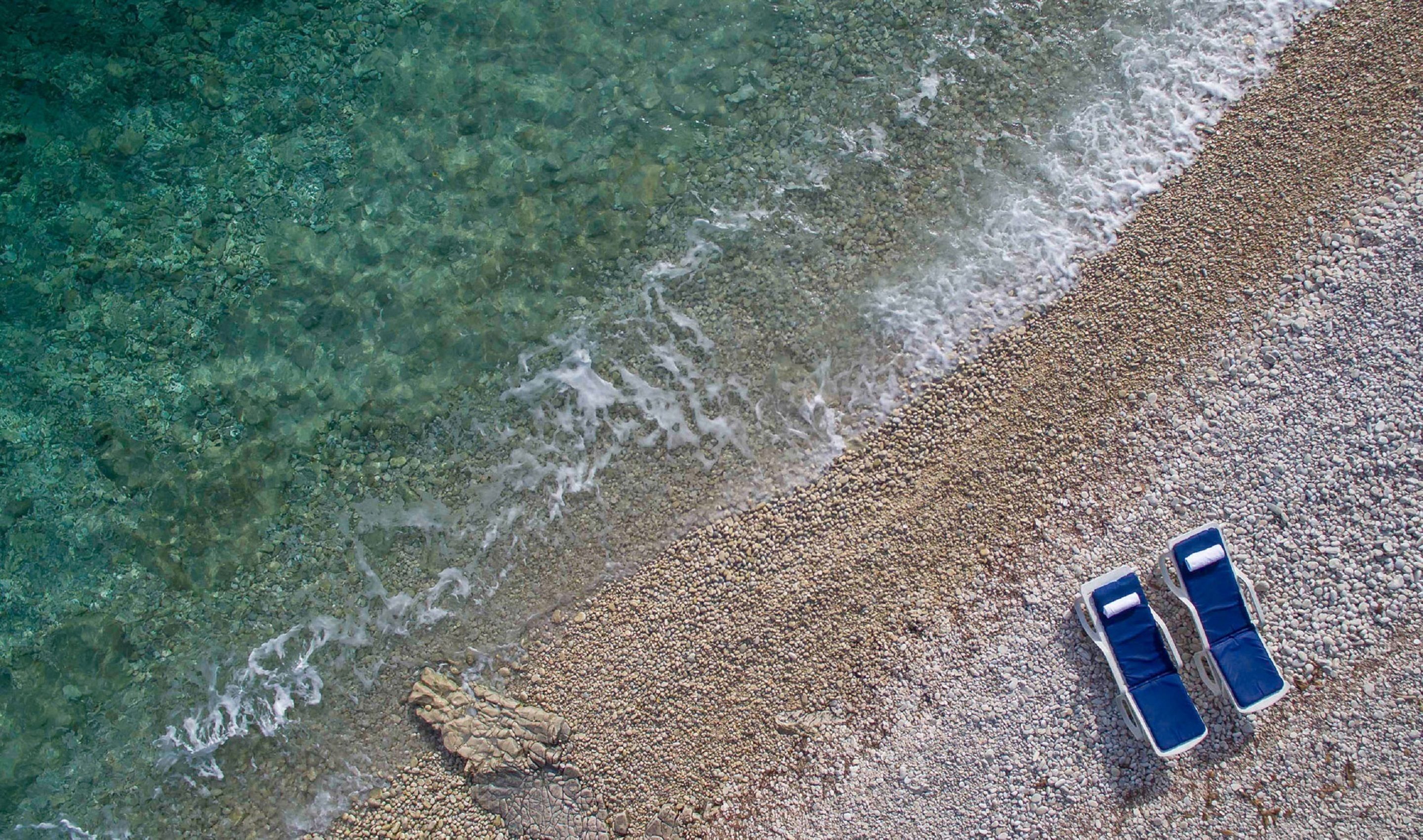 An aerial view of a beach with pebbles and clear water. Two blue lounge chairs are placed on the pebbles.