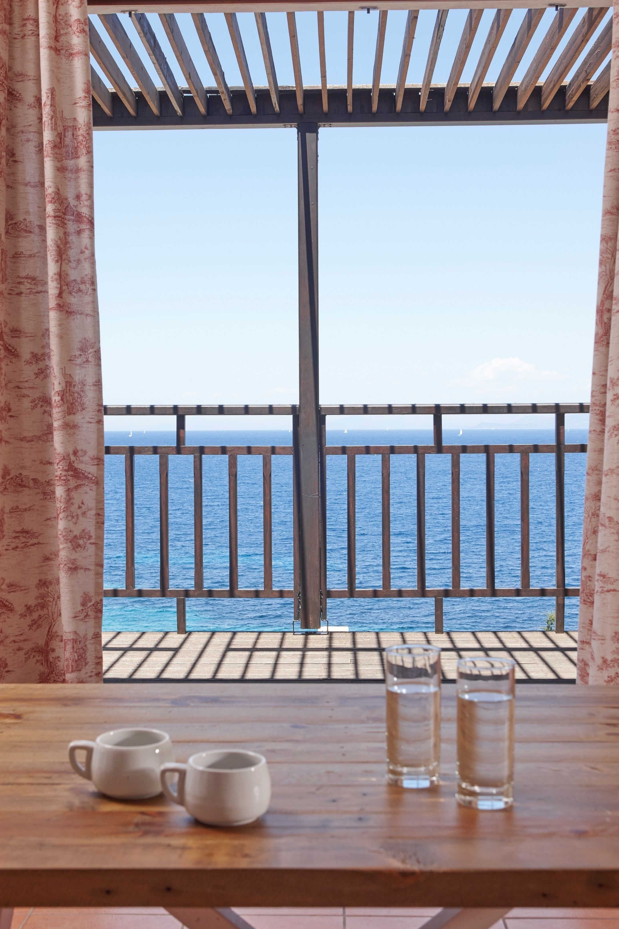 A wooden table with two white mugs and two clear glasses, set against a backdrop of a wooden pergola and a view of the sea through a railing.