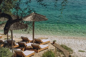 A beachside lounge with wooden lounge chairs and a thatched umbrella. - Anassa Mare Villas & Residences by GHH (Paxos)
