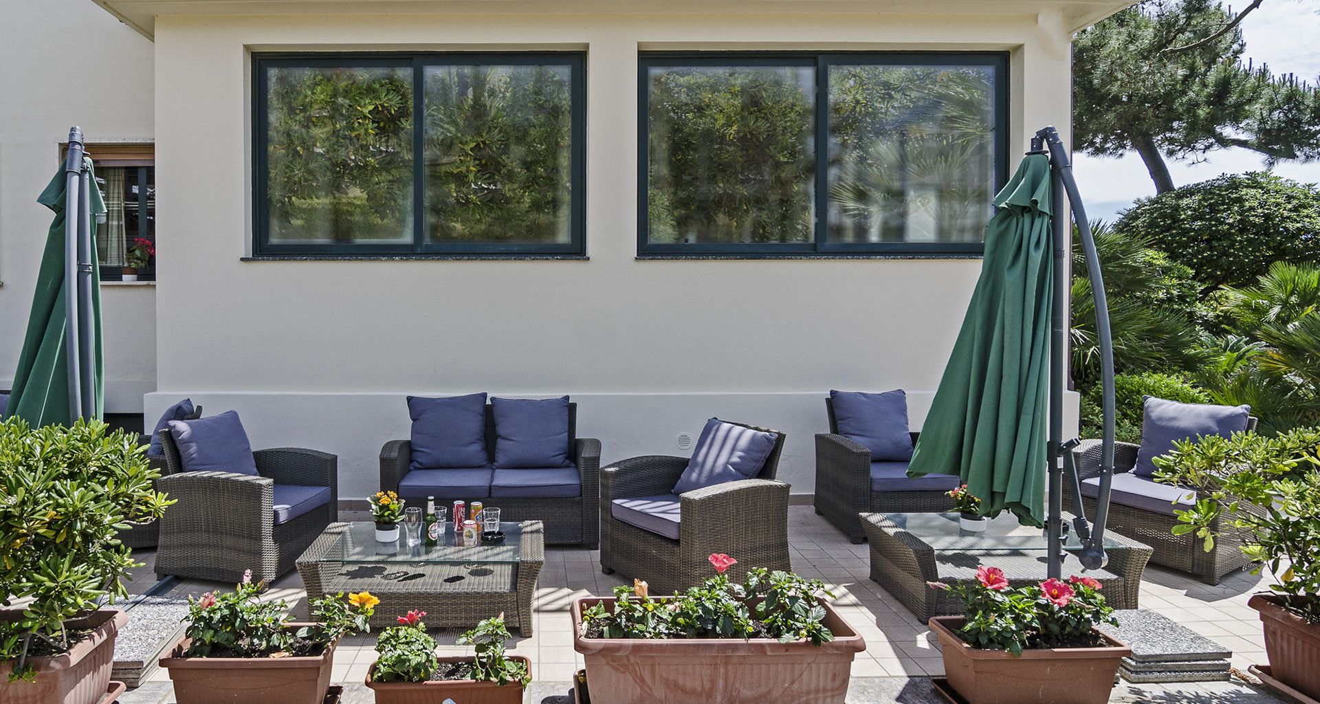An outdoor seating area with wicker furniture and green umbrellas, surrounded by potted plants and a building with large windows.