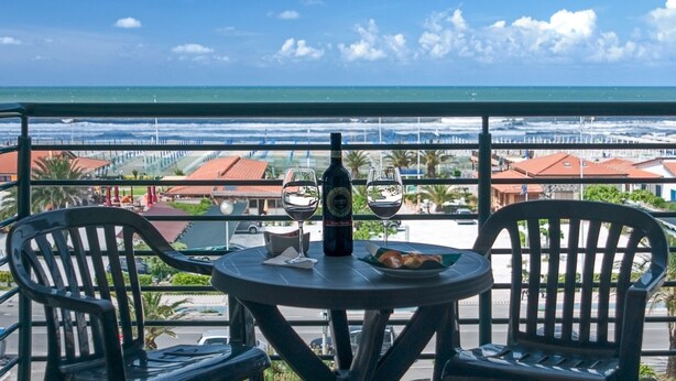 Un balcon avec une table, deux chaises, une bouteille et deux verres. La vue comprend une plage et des bâtiments au loin.