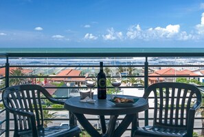 Un balcón con una mesa, dos sillas, una botella y dos vasos. Se ve una playa y edificios a lo lejos.