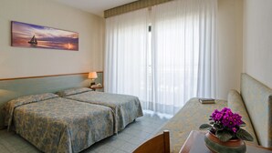 Une chambre d’hôtel avec deux lits, un canapé, une table ornée d’un vase de fleurs, un tableau représentant un voilier et une fenêtre avec des rideaux légers.