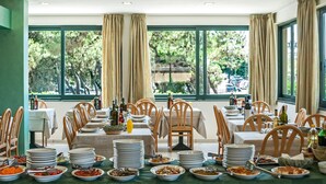 Un espace repas avec plusieurs tables dressées pour un repas, entouré de grandes fenêtres ornées de rideaux, et offrant une vue sur la verdure à l’extérieur.