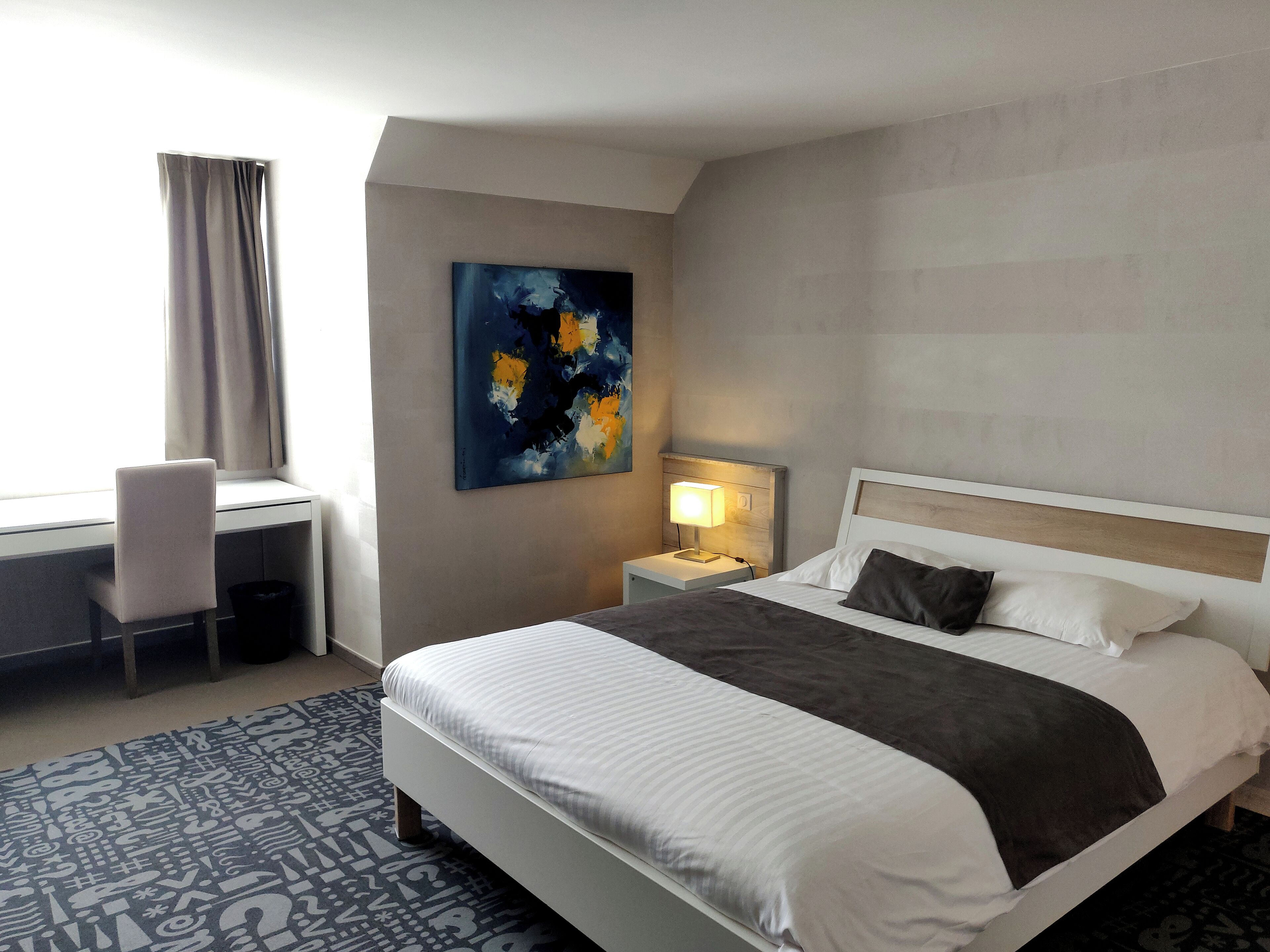 A hotel room with a bed, a desk, a chair, a nightstand with a lamp, and a painting on the wall.