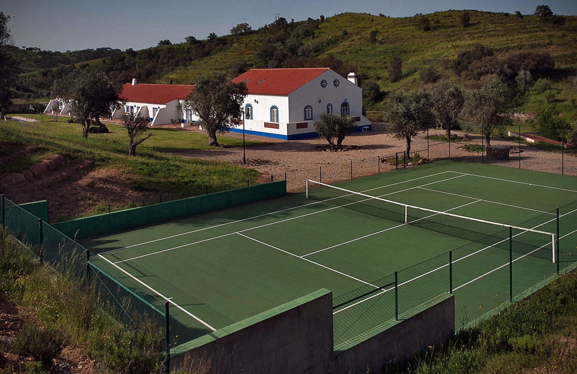 Un campo da tennis con una rete, circondato da una recinzione verde. Sullo sfondo, un edificio bianco con tetto rosso e decorazioni blu.