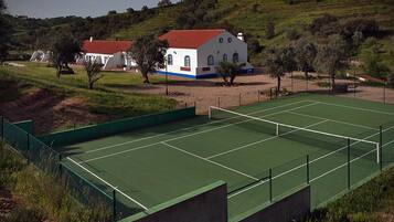 Un campo da tennis con una rete, circondato da una recinzione verde. Sullo sfondo, un edificio bianco con tetto rosso e decorazioni blu.