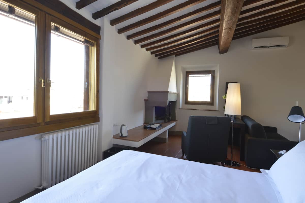 A hotel room with a bed, a desk with a lamp, a chair, and a view of the city through large windows.