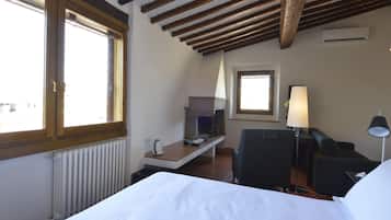 Ein Hotelzimmer mit einem Bett, einem Schreibtisch mit Lampe, einem Sessel und Blick auf die Stadt durch große Fenster.