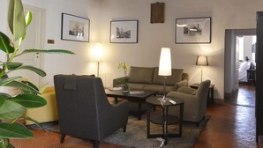Una sala de estar con sofá, sillones, una mesa de centro y una lámpara. En la pared hay cuadros enmarcados y una planta en la esquina.