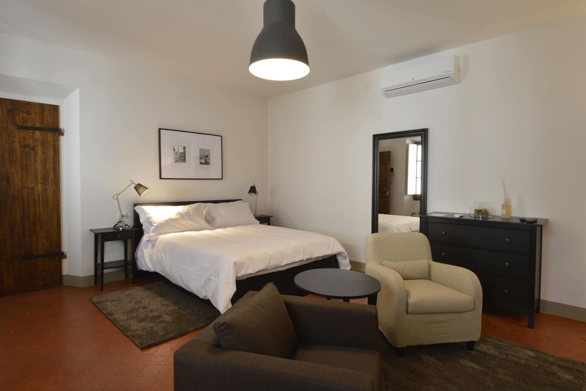 A bedroom with a bed, a chair, a small table, a desk, a mirror, and an air conditioning unit.
