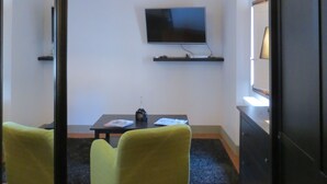 Una habitación con una televisión montada en la pared, una pequeña mesa con una botella y papeles, dos sillones verdes y un mueble negro.